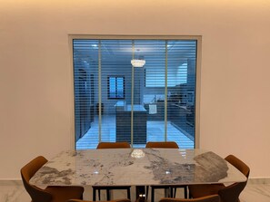 Ein Essbereich mit einem Marmortisch und Stühlen, Blick durch ein Fenster auf eine Küche und einen Essraum, sowie eine Chandelier.