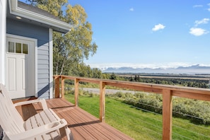 A wooden deck with a white chair, a small building with a door and windows, and a view of a landscape with trees and mountains.