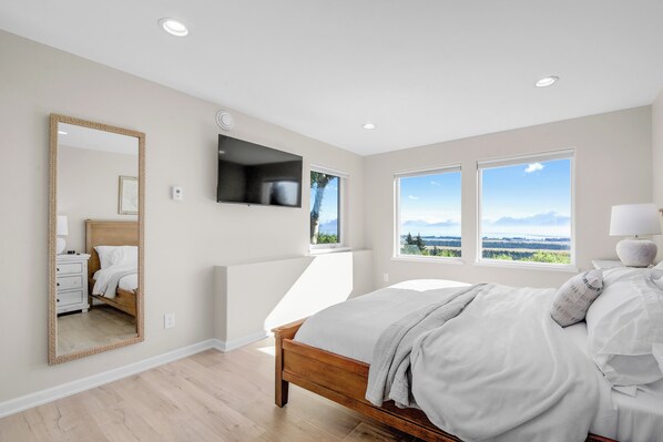 A bedroom with a large bed, a flat-screen TV, a mirror, and two windows offering a mountain view.