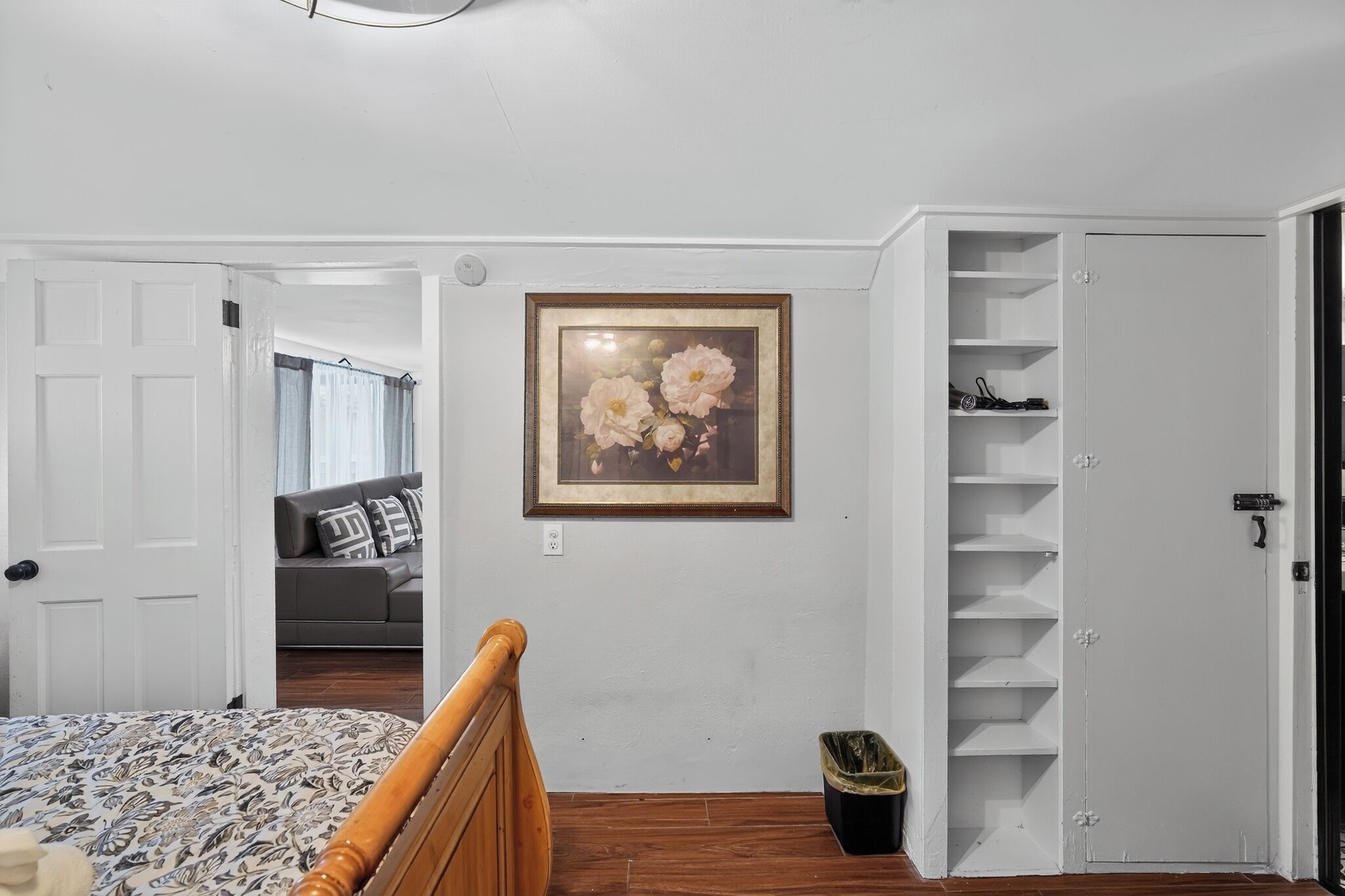 A bedroom with a bed, a wooden headboard, a framed floral artwork on the wall, a white door, and a closet with shelves.