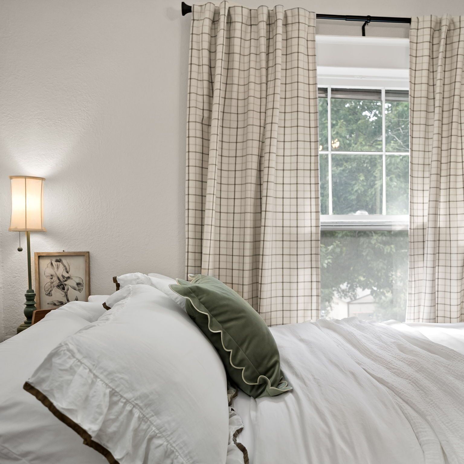 Een slaapkamer met een bed, witte beddengoed, een groen kussen, een nachtkastje met een lamp en een raam met gordijnen.