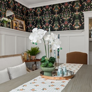 A dining area with a table set for two, featuring a floral table runner and a tray with a green plant centerpiece.