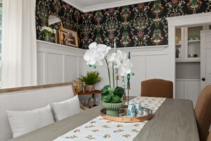 Een eetgedeelte met een tafel gedekt voor twee personen, bloemenbehang en een wit kastje op de achtergrond.