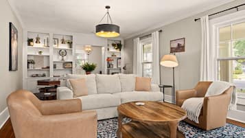 Ett vardagsrum med en soffa, två fåtöljer, ett soffbord och en golvlampa. Det finns hyllor med dekorativa föremål och ett fönster med gardiner.
