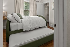 A single bed with a green headboard and white bedding in a room with white walls and a window with curtains.