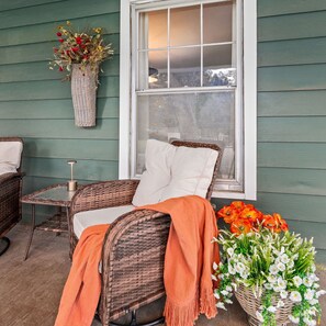 A wicker chair with a white cushion and an orange throw blanket, a small table with a candle, and a window with a view of the outdoors.