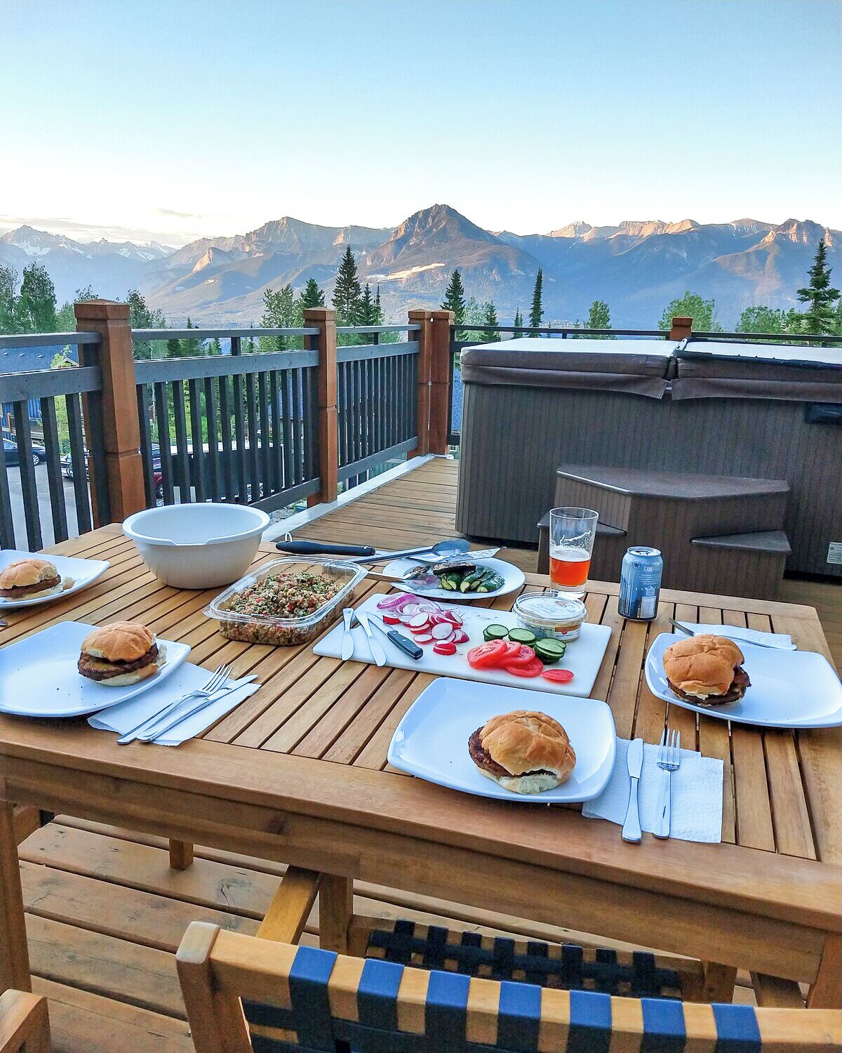 Eine Holzterrasse mit Tisch, gedeckt für eine Mahlzeit, mit Burgern, Salaten und Getränken, mit Blick auf eine gebirgige Landschaft.