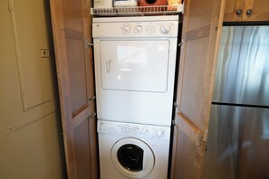Un lavadero pequeño con lavadora y secadora apiladas, armarios de madera y un refrigerador de acero inoxidable.