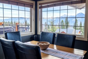 A dining area with a table and chairs, a wooden bowl, and a view of mountains through large windows.