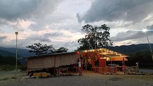 A rustic outdoor dining area with a covered seating area and a small building, surrounded by natural scenery.