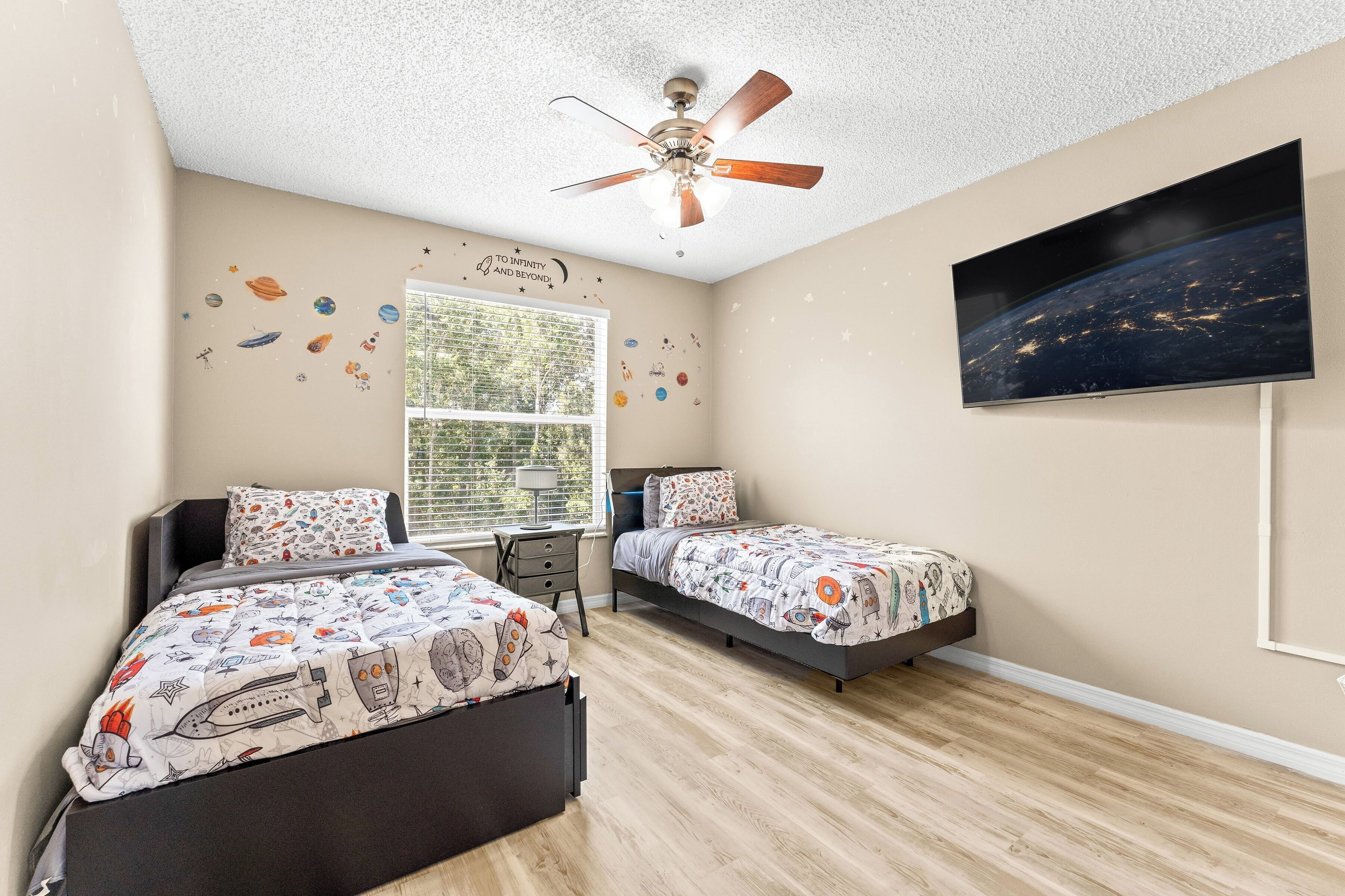 A bedroom with two beds, a ceiling fan, a window with blinds, and a wall-mounted TV.