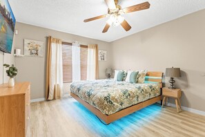 Ein Schlafzimmer mit einem Bett, Deckenventilator, Nachttisch, Lampe und einem Fenster mit Vorhängen.