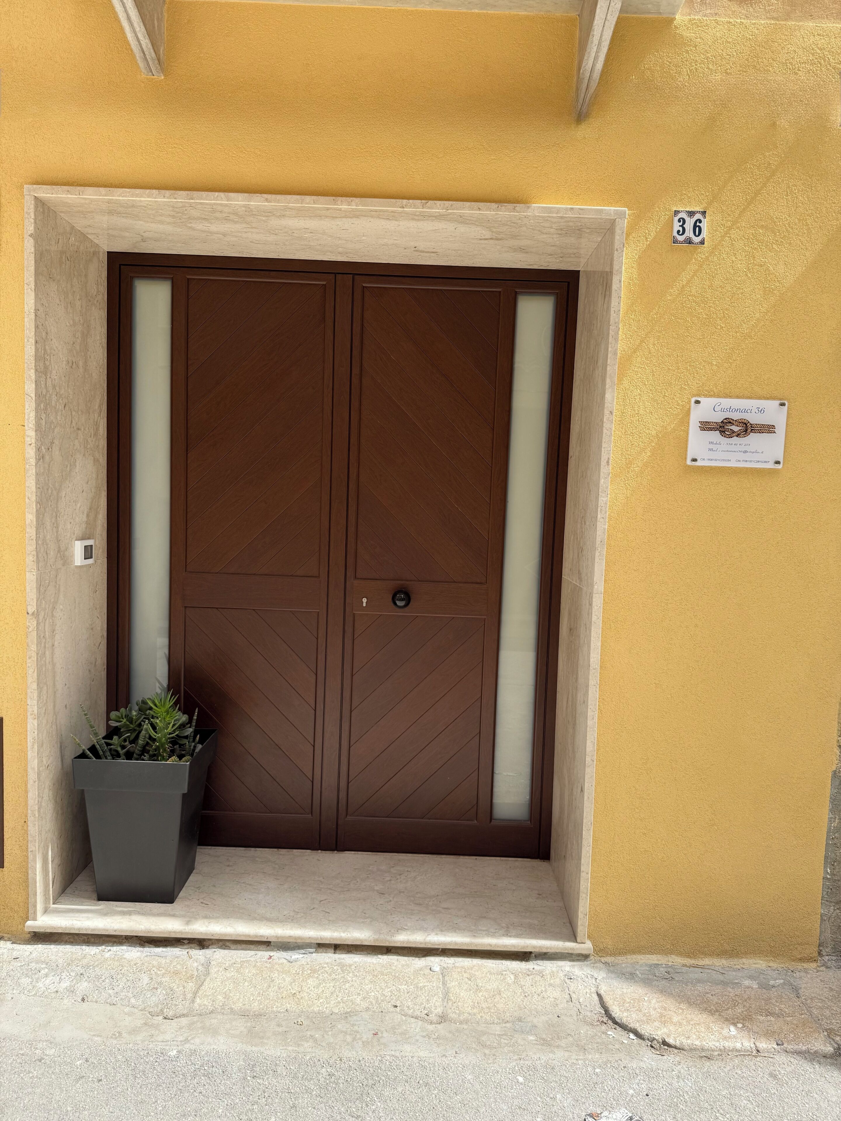 A double wooden door with a glass panel, a potted plant, and a sign with the number 36 on a yellow wall.