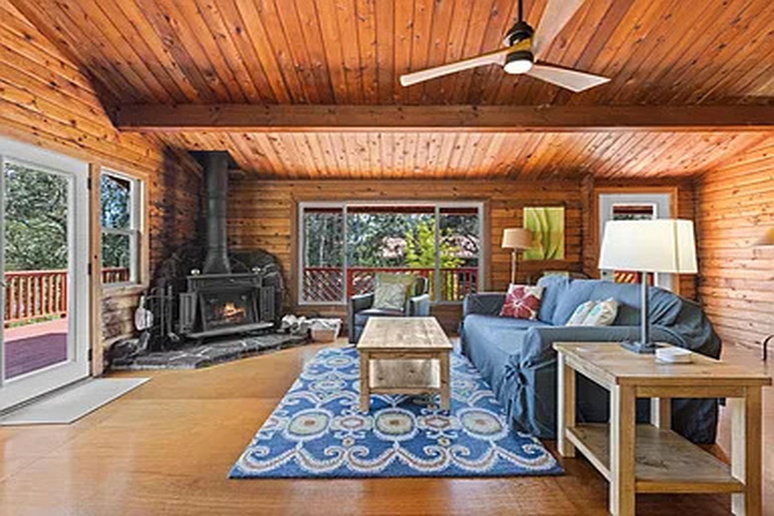 A cozy cabin interior with a fireplace, wooden walls, a sofa, and a ceiling fan.