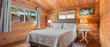 Ein Schlafzimmer einer Holzhütte mit einem Bett, Nachttischen, einem Fenster und Blick ins Freie.