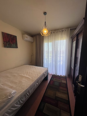 Une chambre avec un lit simple, un luminaire suspendu, un tableau au mur et une fenêtre avec des rideaux.