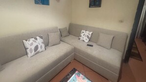 A corner living room with a grey sofa, two patterned cushions, a remote control on the sofa, and a small decorative wall hanging. - Perfect chalet for rent North coast porto golf marina (Marsa Matrouh Governorate)