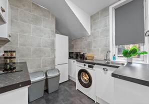 A modern kitchen with grey tiles, a built-in washing machine, a sink, and a window with a plant. - Modern ground floor apartment close to train station (Coatbridge)
