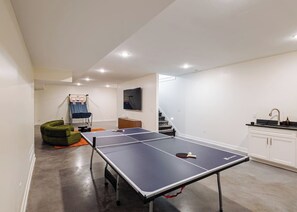 Une table de ping-pong dans une pièce où se trouvent un panier de basket, un téléviseur et un escalier.