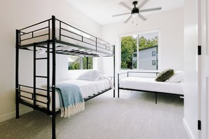 Una habitación con una cama litera, una cama individual, un ventilador de techo y una ventana con vista a una casa.