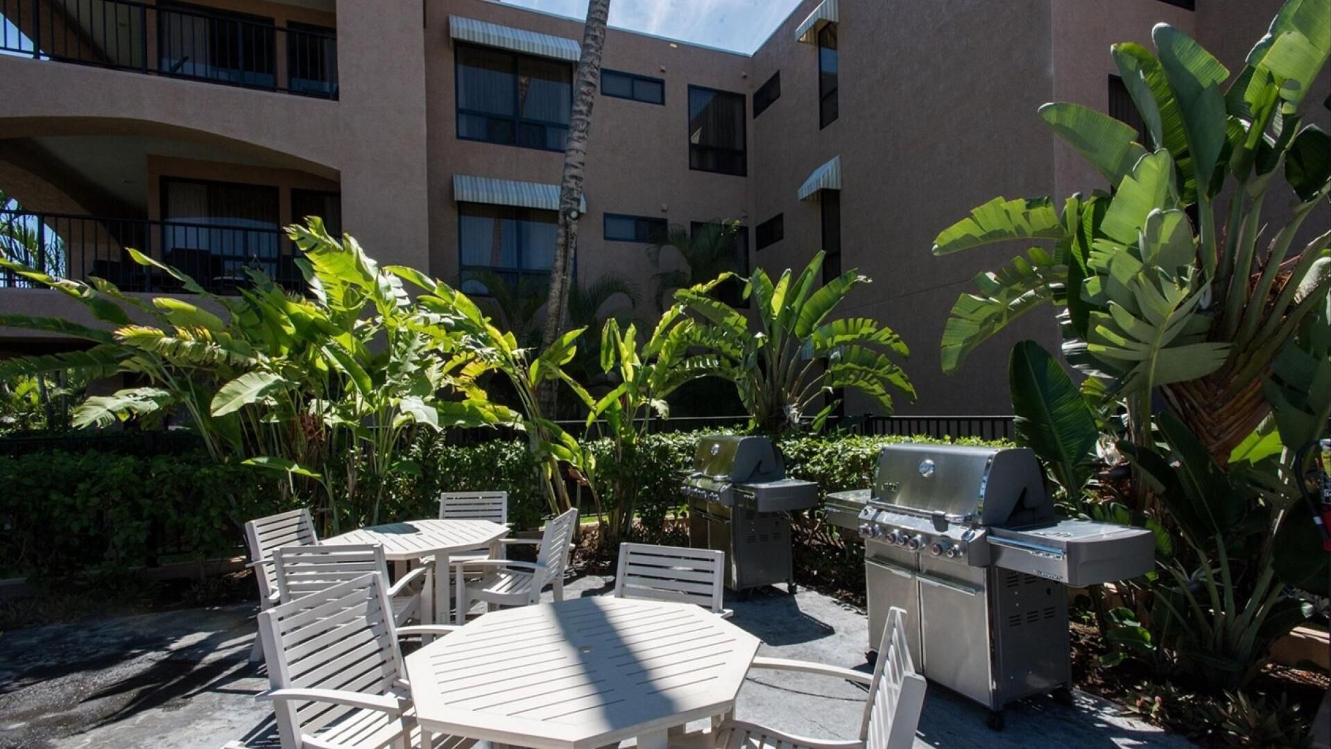 An outdoor patio with white tables and chairs, surrounded by lush tropical plants and a barbecue grill.