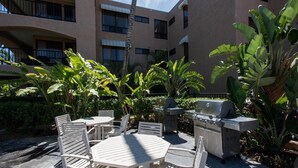 Une terrasse extérieure avec des tables et des chaises blanches, entourée d’une végétation tropicale luxuriante et agrémentée d’un barbecue.