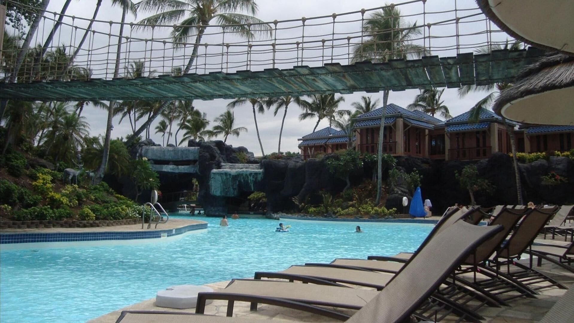 Ein Poolbereich mit Liegestühlen, einer Wasserrutsche und tropischer Umgebung mit Palmen und einem Gebäude im Hintergrund.