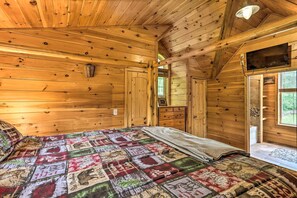 A cabin interior with a wooden ceiling, a bed with a patterned quilt, a window with a view, and a mounted television.