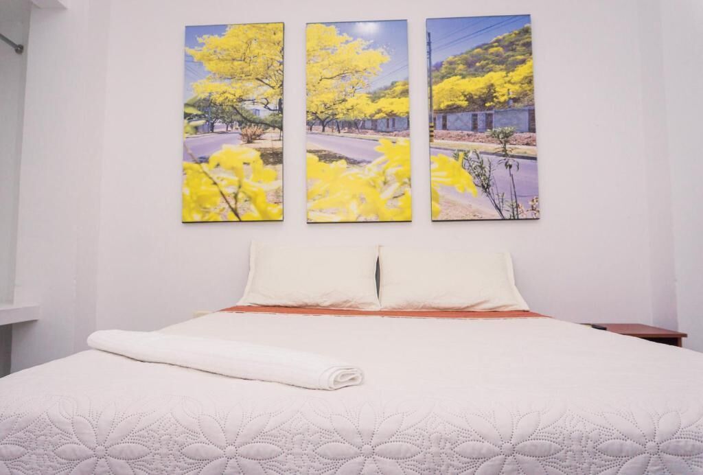 Een bed met een witte deken en kussens, een opgevouwen witte handdoek en een muur met drie gele bloemschilderijen.