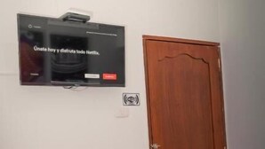 Una habitación con un televisor montado en la pared, una puerta de madera y una mesa con un tapete enrollado.