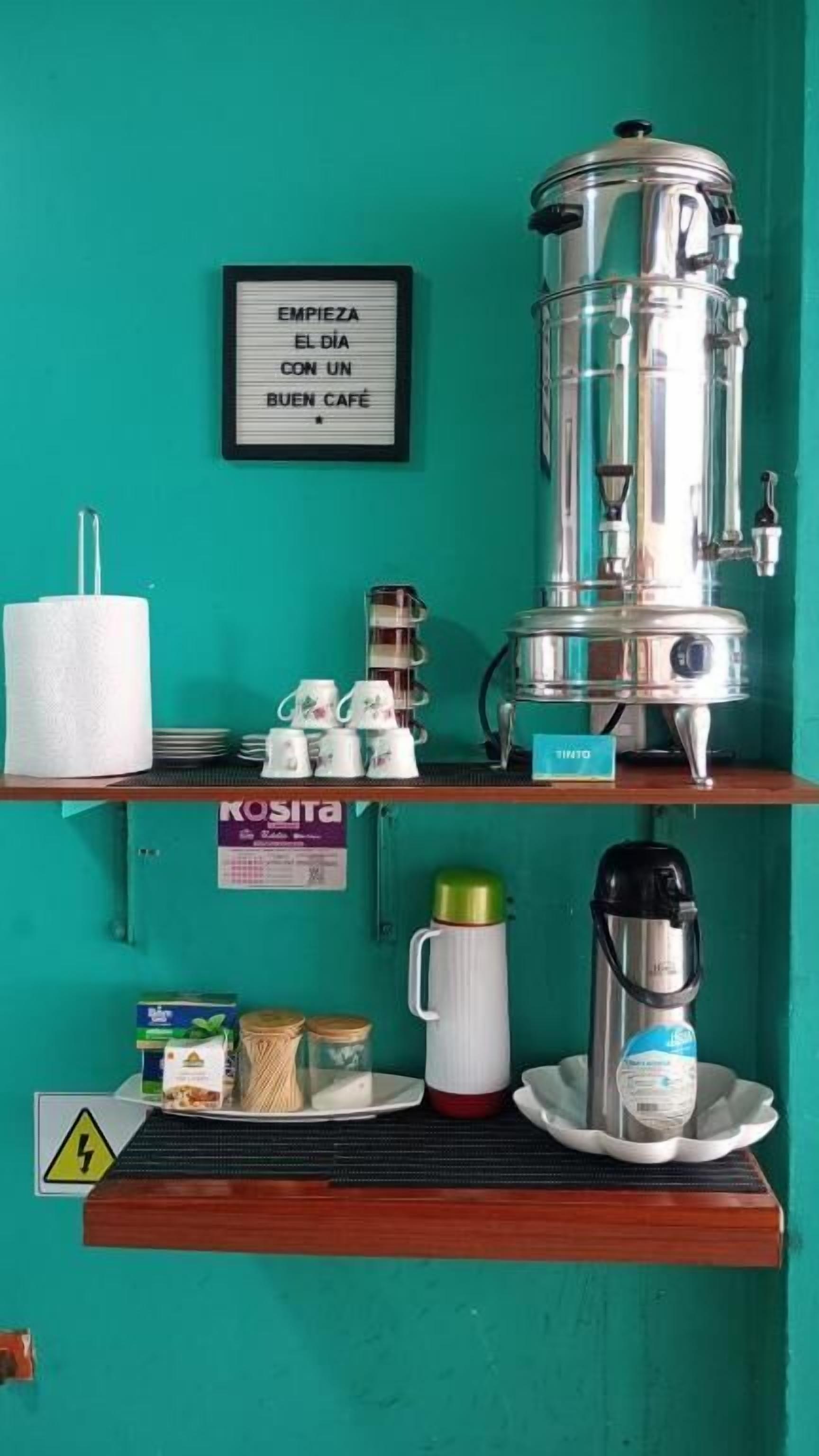Una estación de café con cafetera, tazas y un cartel sobre una pared azul verdoso.
