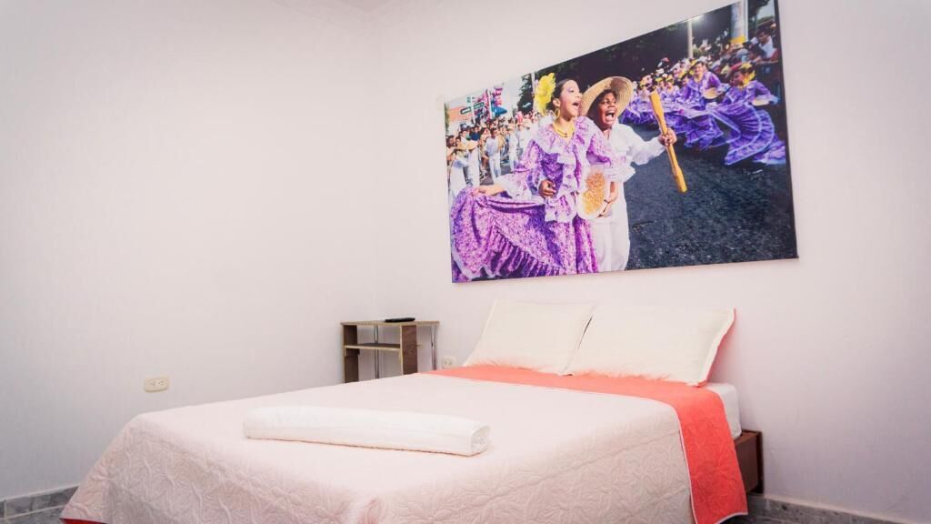 Una habitación de hotel con una cama, una mesita de noche y un cuadro en la pared que muestra un evento cultural.