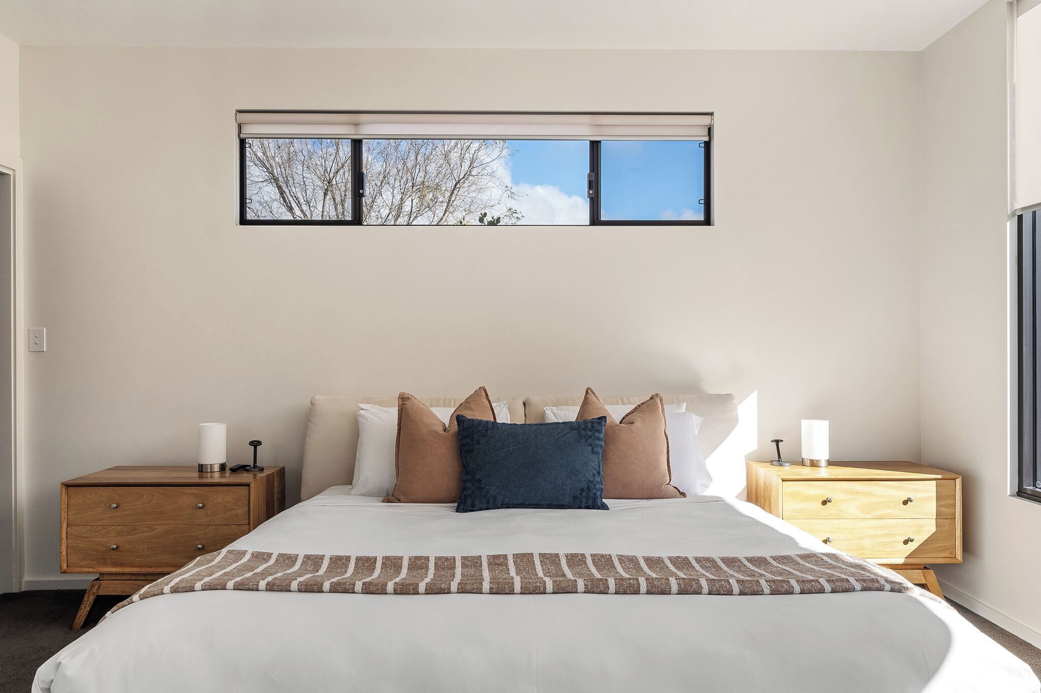 A bedroom with a large bed, two nightstands, a window with blinds, and a view of trees.