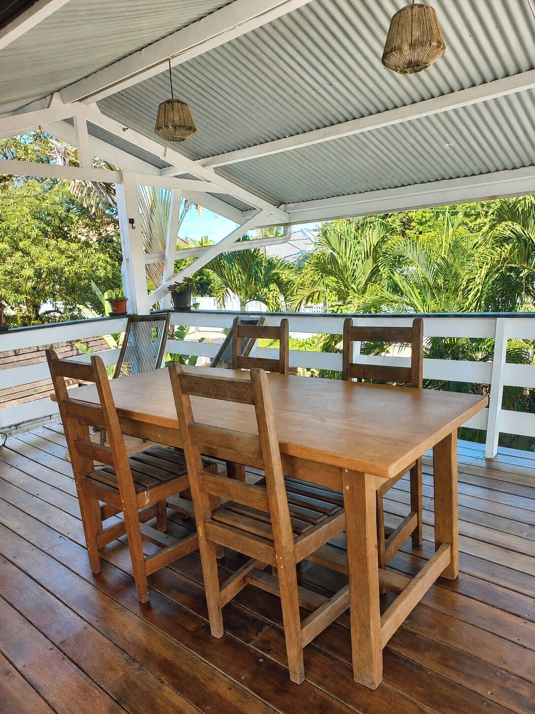Une table à manger en bois, entourée de chaises, placée sous un toit en tôle ondulée, le tout au milieu d’une végétation luxuriante.