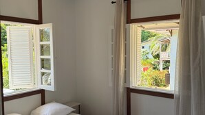 Un dormitorio con piso de madera, una cama con ropa de cama blanca, una lámpara colgante y un ventanal con cortinas.