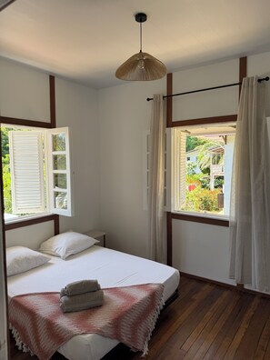 Une chambre avec un plancher en bois, un lit avec une literie blanche, un luminaire suspendu et une fenêtre avec des rideaux.