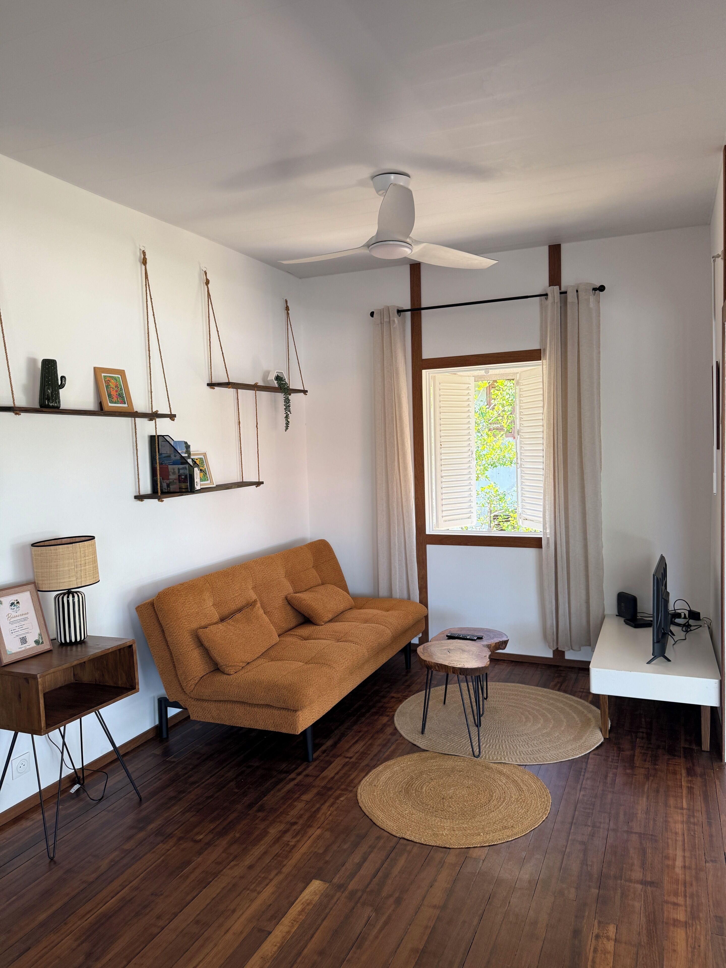 Ein modernes Wohnzimmer mit einer Couch, einem Deckenventilator, einem Fernseher und einem Fenster mit Vorhängen.