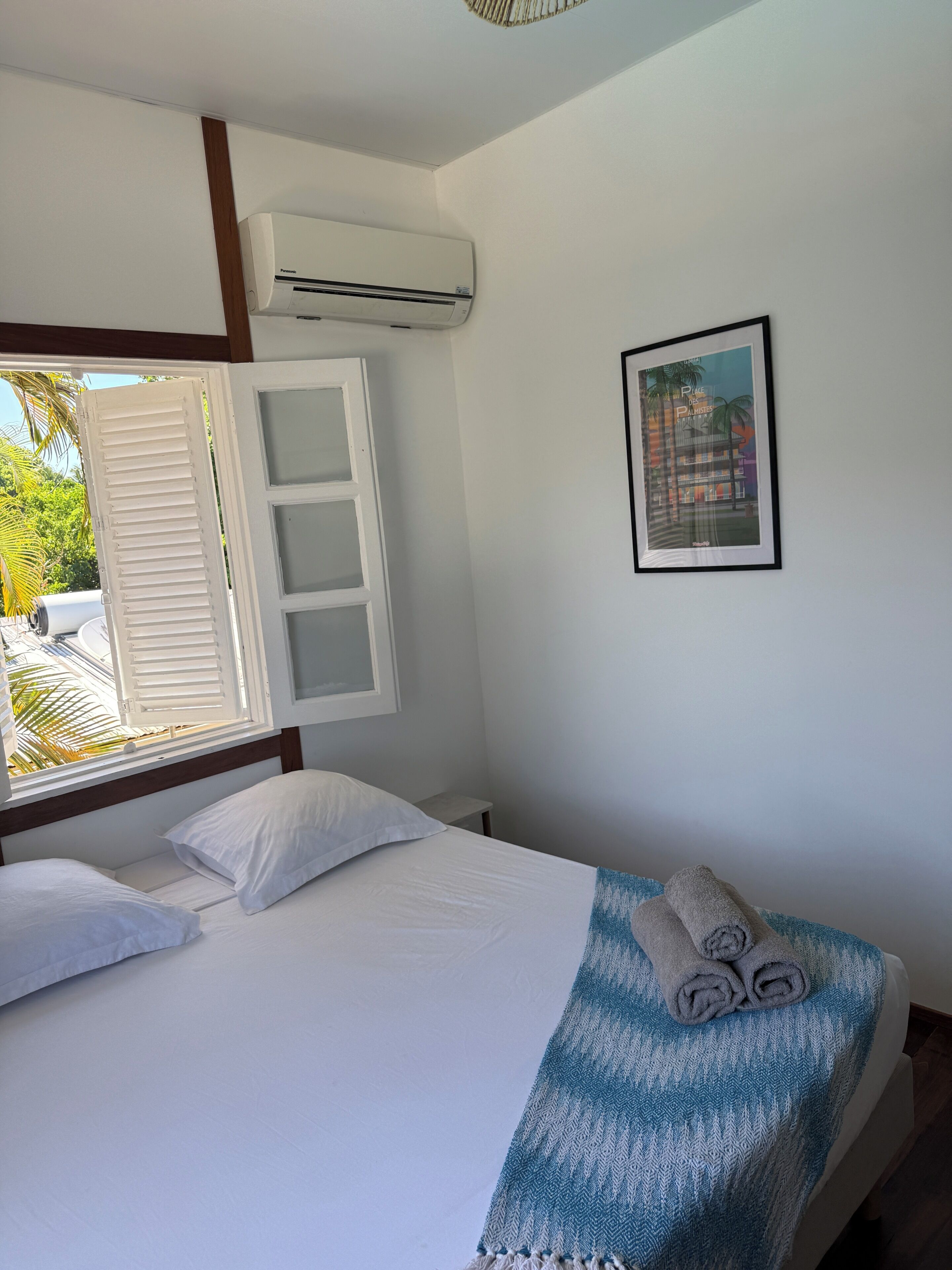 A bedroom with a bed, a window with shutters, an air conditioning unit, a framed picture on the wall, and a towel on the bed.