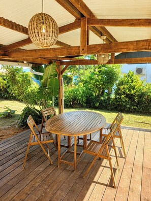 Une terrasse extérieure en bois, avec une table ronde et des chaises, entourée d’une végétation luxuriante.
