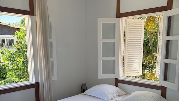 Un dormitorio con cama, cortinas blancas, una ventana con contraventanas y vistas al exterior.