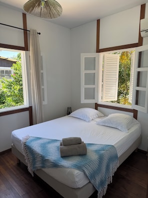 A bedroom with a bed, white curtains, a window with shutters, and a view of greenery outside. - Kaz Toti - Private pool - Wi-Fi - Air conditioning - Beach 100 m away - Montabo - Cayenne (Cayenne)