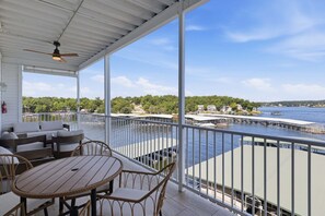 A covered outdoor area with a table and chairs, a ceiling fan, and a view of a body of water with docks and buildings in the distance. - Main-channel Views, Heated Pool, Boat Slips & Huge Deck (Osage Beach)