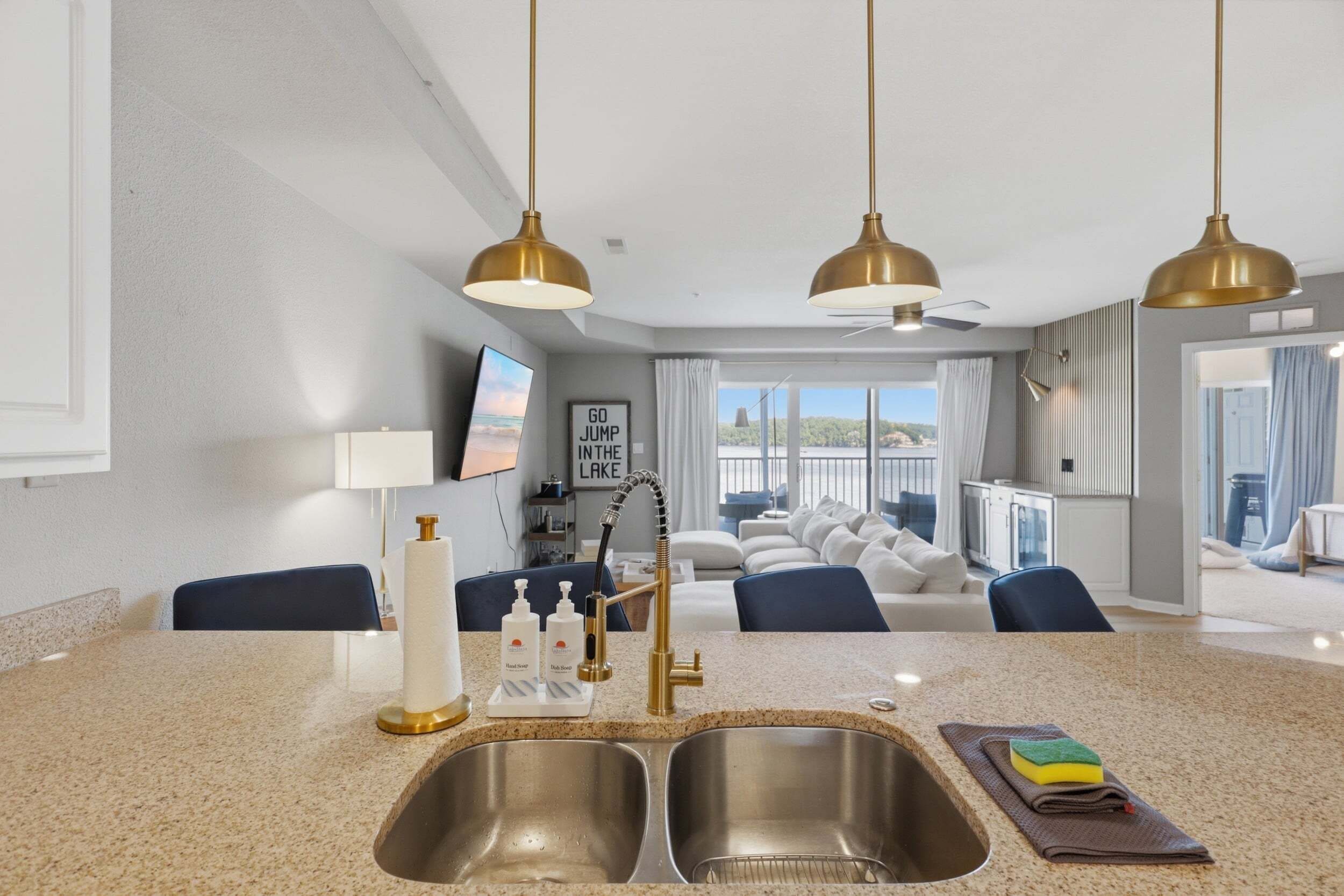 A modern kitchen with a double sink, a dining area with a view, and a living room with a sofa and a ceiling fan.