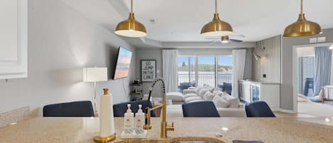 A modern kitchen with a double sink, a dining area with a view, and a living room with a sofa and a ceiling fan.