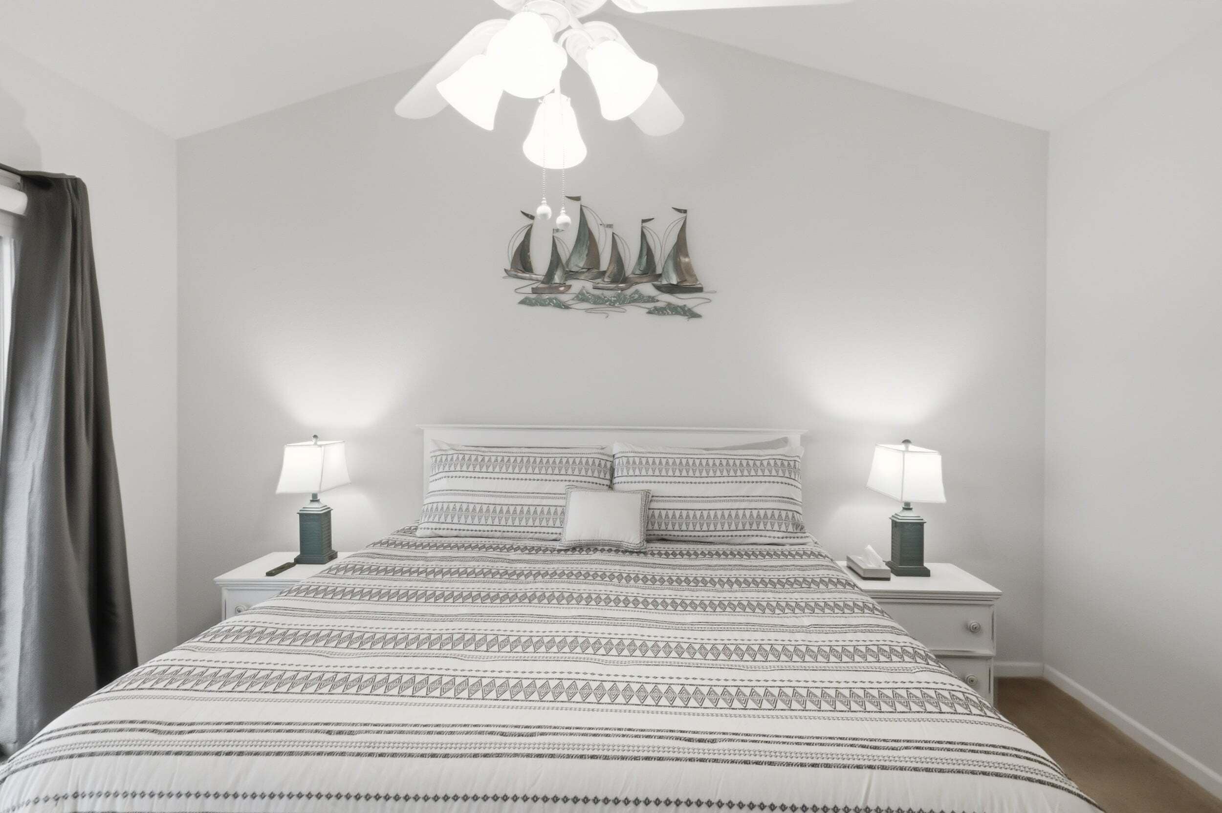 A bedroom with a bed, two bedside tables with lamps, a ceiling fan, and a wall decoration of sailboats.