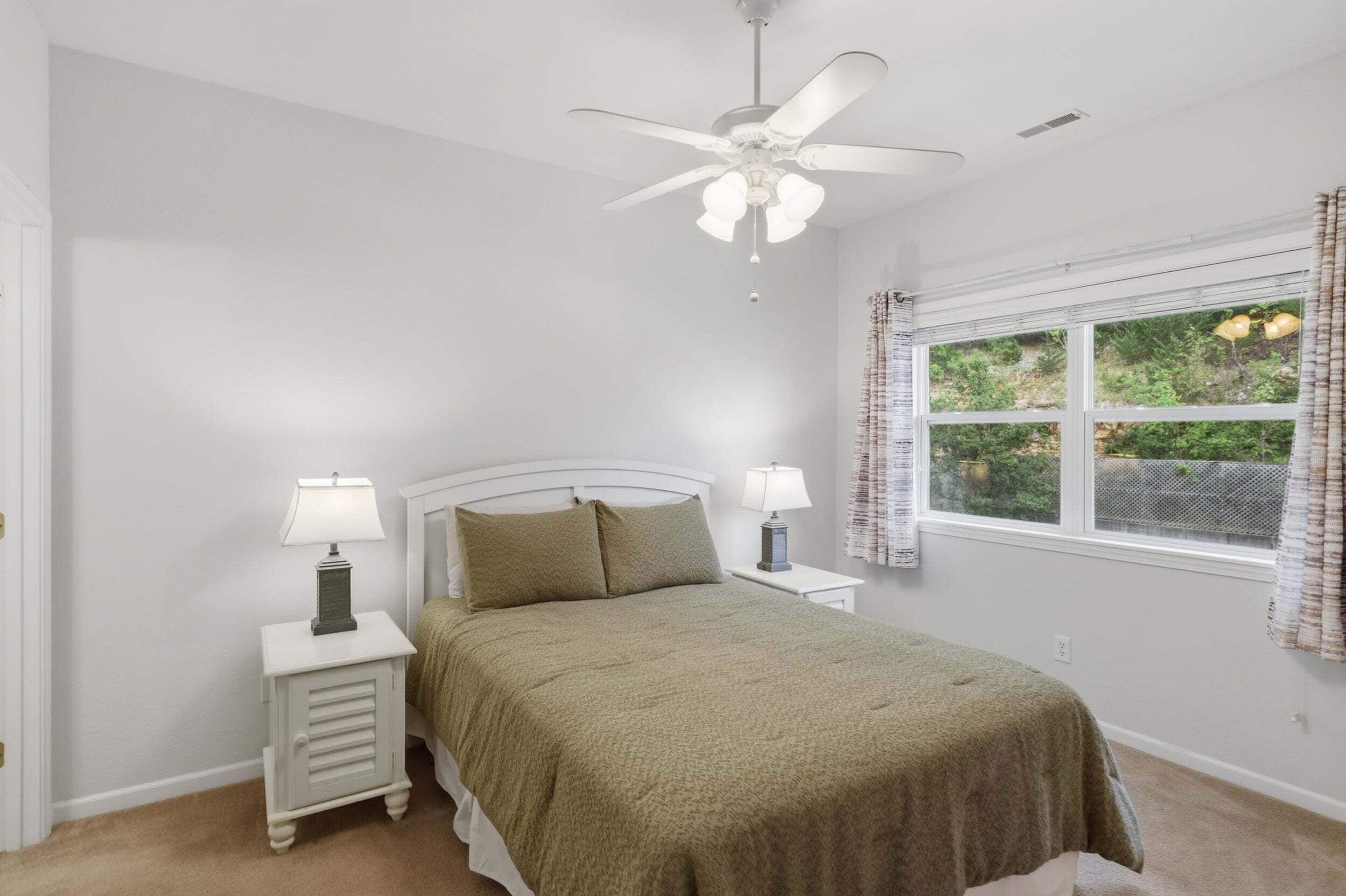 A bedroom with a bed, two bedside tables with lamps, a ceiling fan, a window with curtains, and a view of greenery outside.