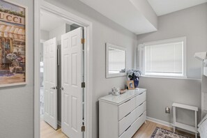 Une pièce avec une commode blanche, un miroir, un vase de fleurs et un tableau encadré posé sur la commode.