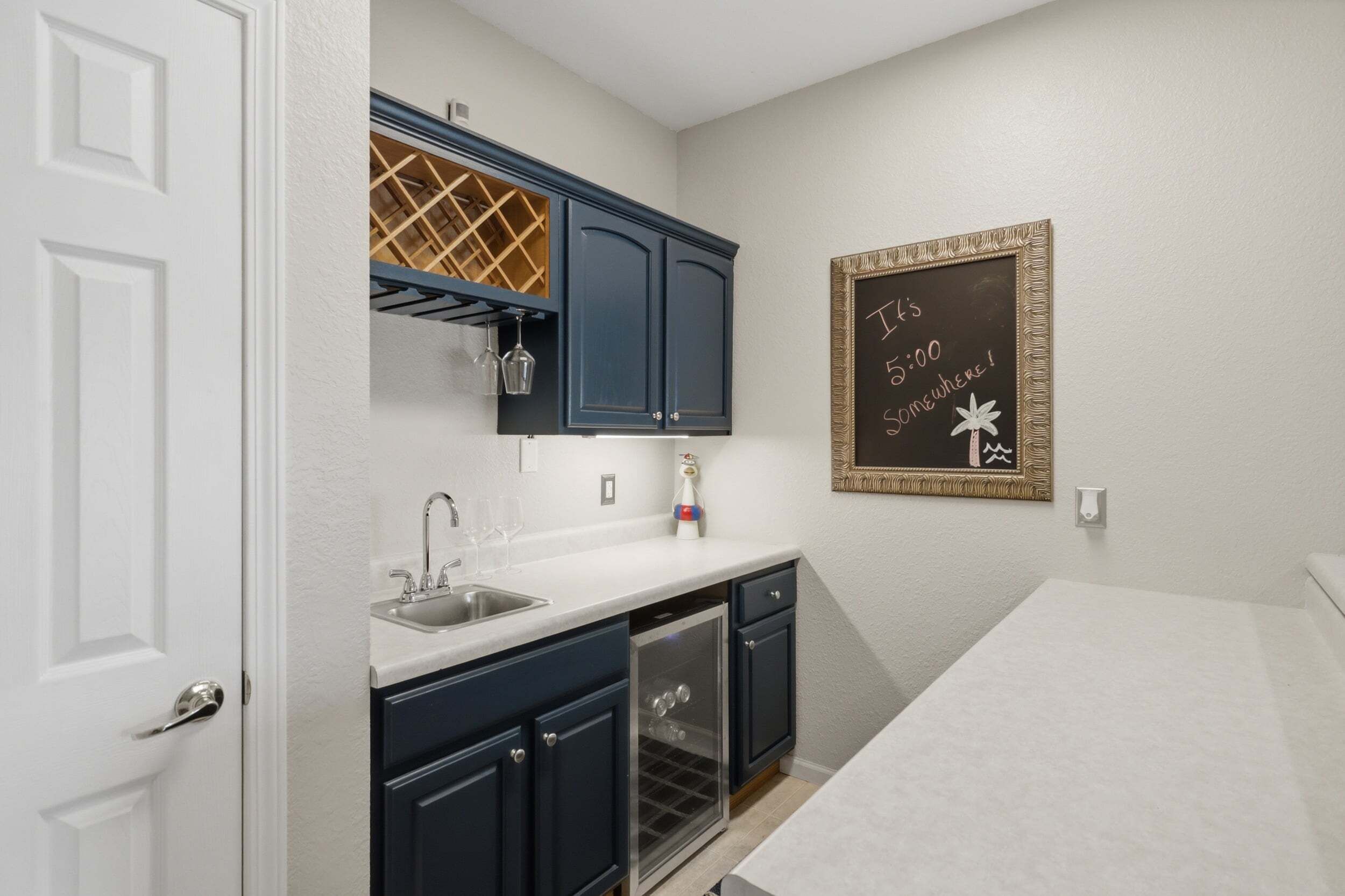 A kitchen with a white countertop, dark cabinets, and a wine rack. There's a chalkboard with writing on the wall.