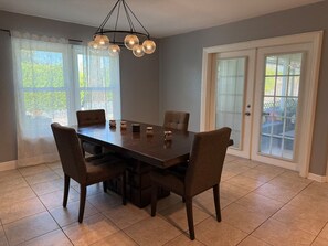 Un comedor con una mesa de madera oscura y cuatro sillas, una araña con múltiples lámparas y vistas al exterior a través de cortinas translúcidas.