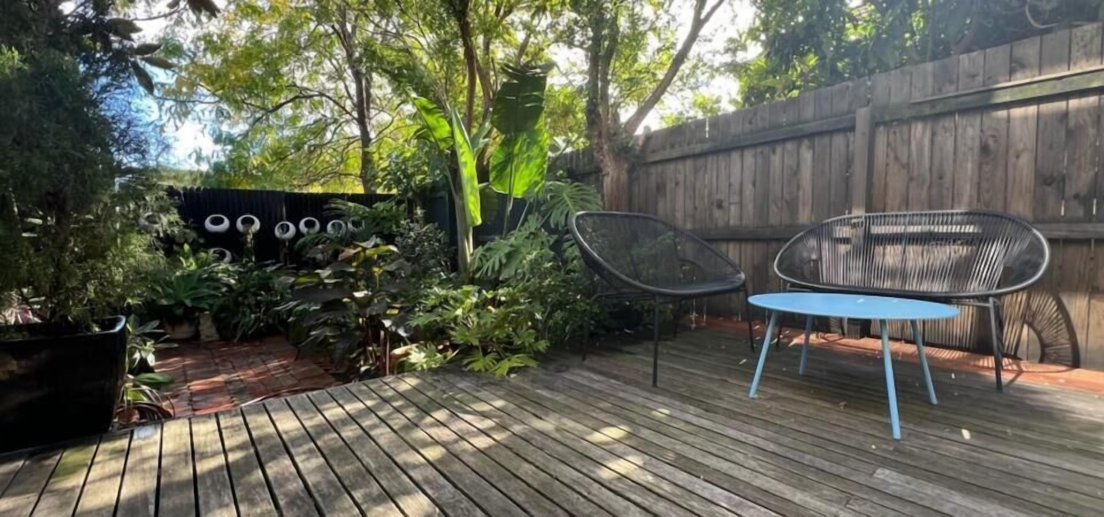 Una terraza de madera con dos sillas de metal y una mesa, rodeada de abundante vegetación y una cerca de madera.
